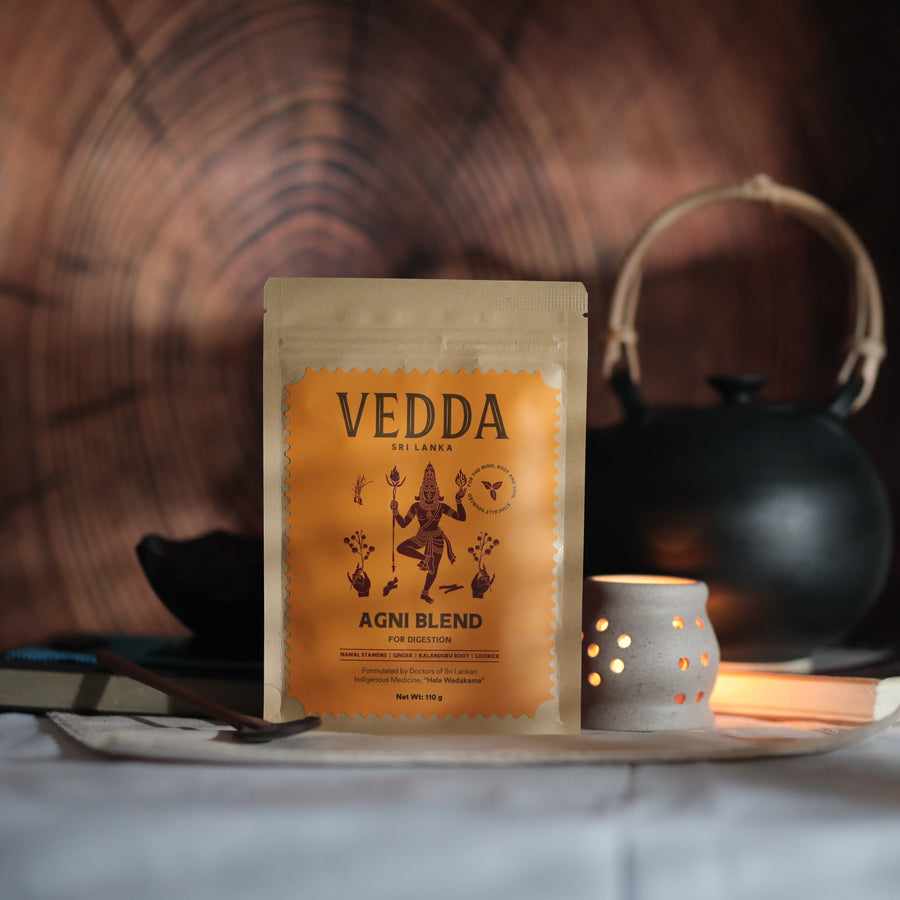 A stand-up pouch of VEDDA AYURVEDA Agni Blend powder sits on a tray in a styled setting with a black teapot, a lit candle holder, and a wood grain background.