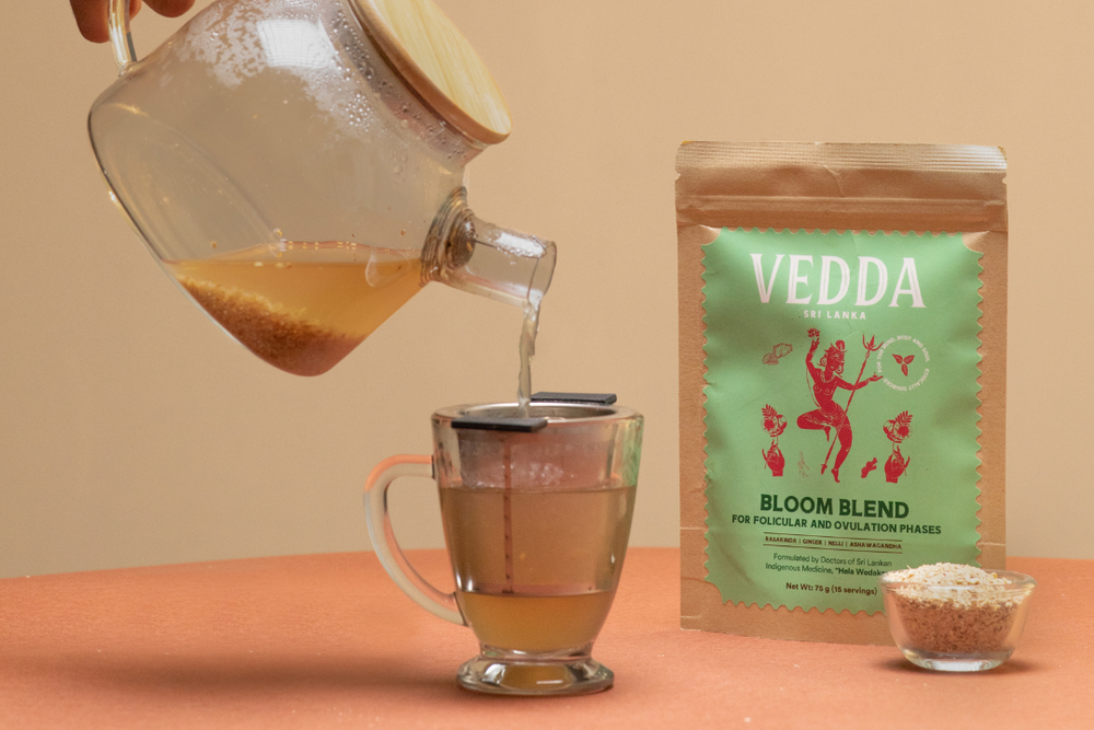 A hand pours tea from a glass teapot with a wooden lid into a glass mug with a metal filter, with a bag of VEDDA Tea 'Bloom Blend' and a small bowl of loose tea on a terracotta surface against a beige background.
