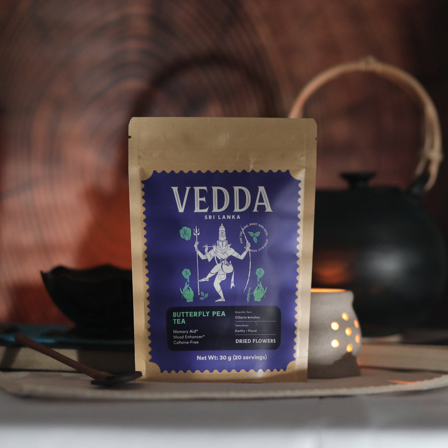A stand-up pouch of VEDDA Kelaka Butterfly Tea Tea centered on a table, with a dark teapot and a small glowing candle in the blurred background against a warm wooden surface.