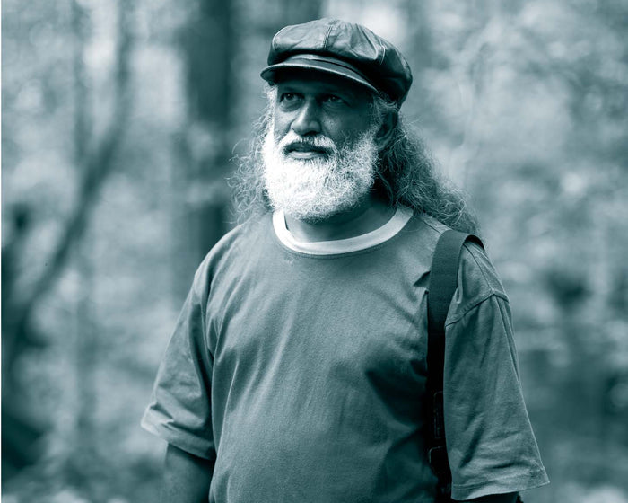 A black and white portrait of a man from the Vedda Tribe with a long white beard wearing a flat cap, t-shirt, and shorts, standing outdoors in a wooded setting.