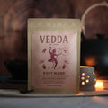 A stand-up pouch of VEDDA Root Blend tea placed on a tabletop in a warm, moody setting with a blurred wooden background and a small glowing candle holder.