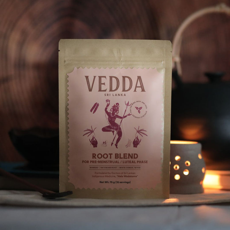 A stand-up pouch of VEDDA Root Blend tea placed on a tabletop in a warm, moody setting with a blurred wooden background and a small glowing candle holder.