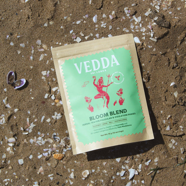 A kraft paper pouch of VEDDA Tea  'Bloom Blend' herbal tea lies on sandy ground scattered with small white shells. The pouch has a light green label with a red dancer illustration and black text.