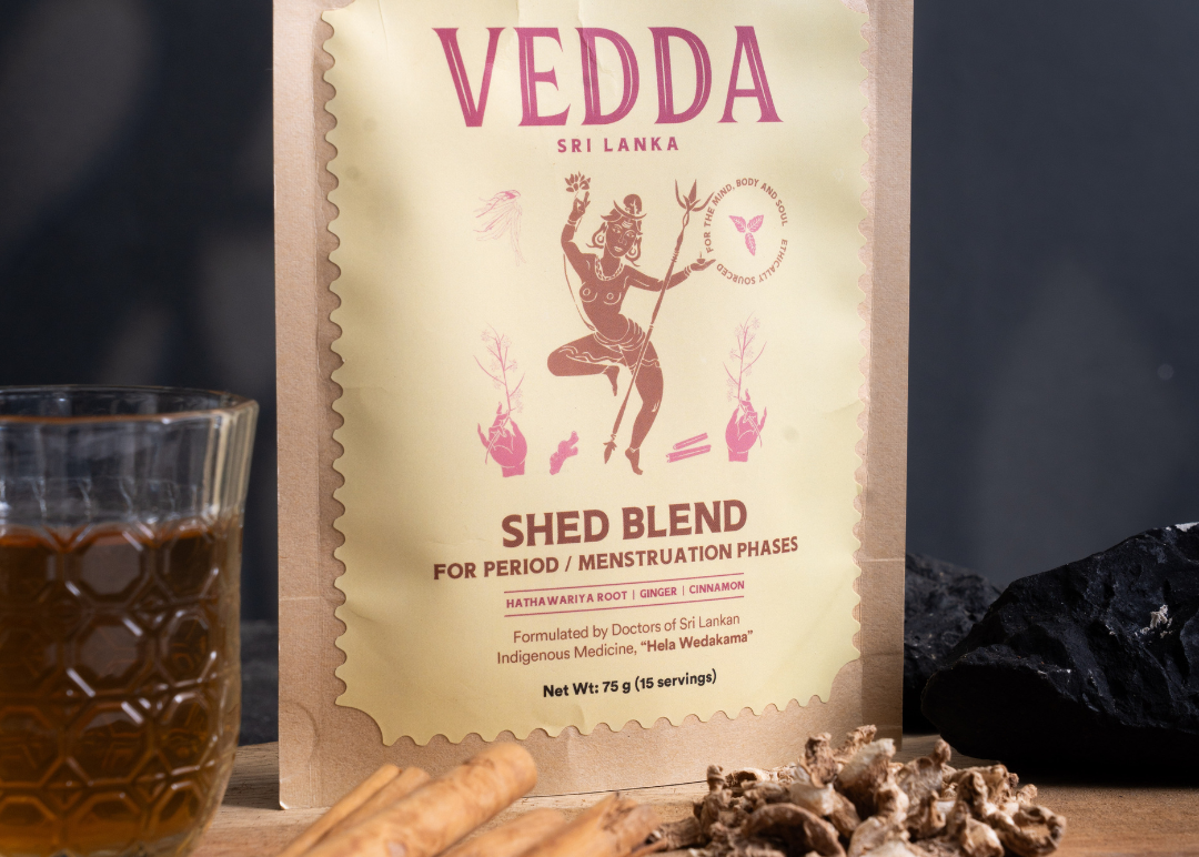 A stand-up pouch of VEDDA Sri Lanka 'Shed Blend' herbal tea is displayed on a wooden surface with cinnamon sticks, dried roots, and a glass of amber liquid against a dark background.