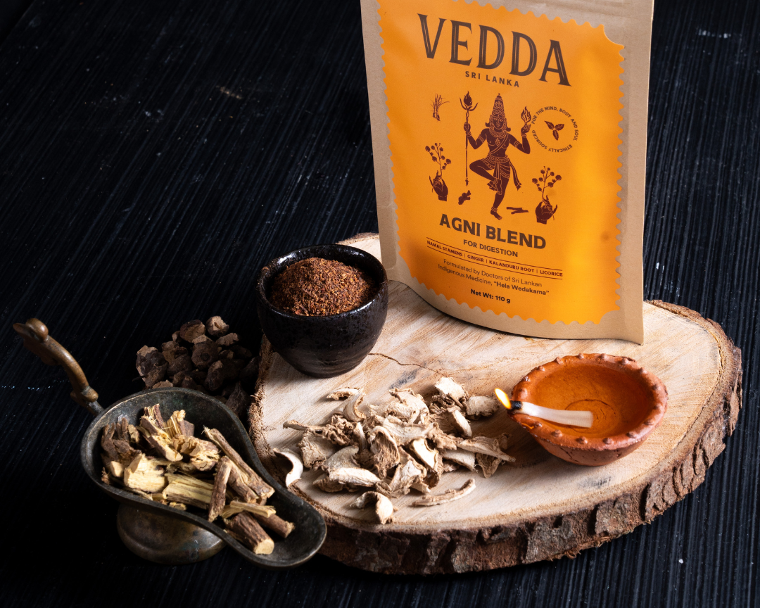 A stand-up pouch of VEDDA Agni Blend (spice infusion) displayed on a rustic wood slice with its ingredients (root herbs, powder, and a clay pot) on a dark wooden table.